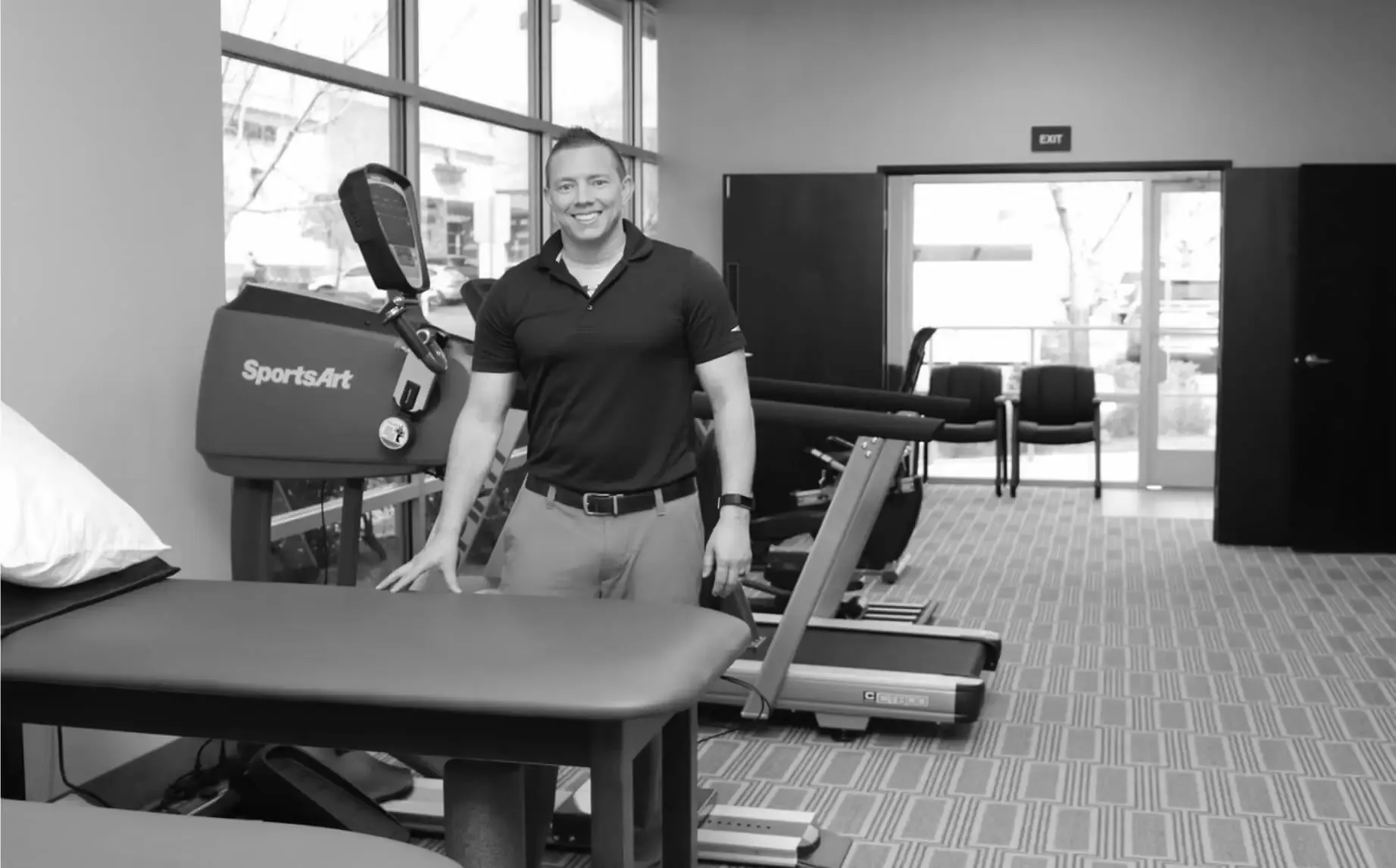 A smiling man in a polo shirt stands by a treatment table in a bright physical therapy gym with treadmills and tall windows