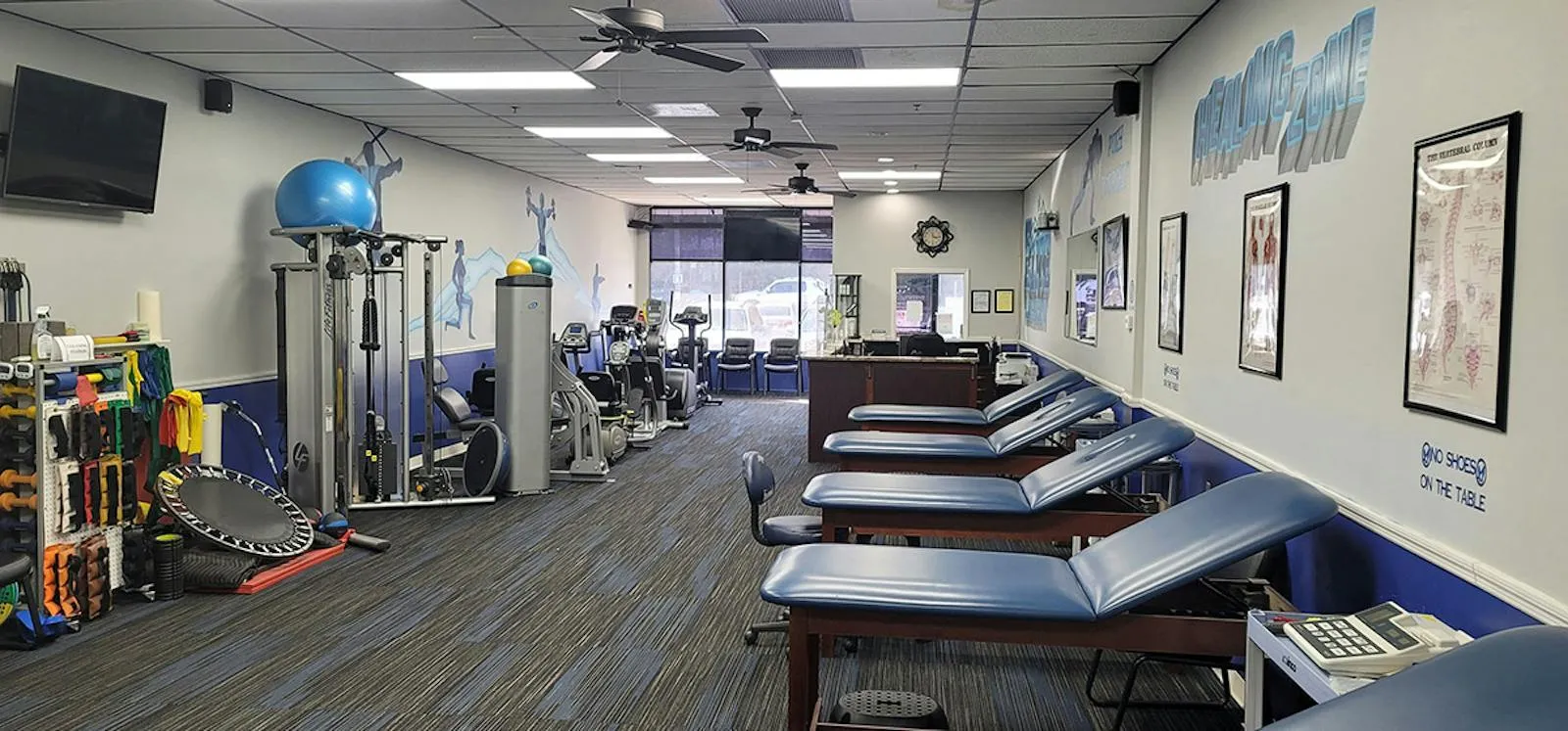 A row of blue treatment tables along a wall painted “Healing Zone”, with exercise equipment and an exercise ball across the room